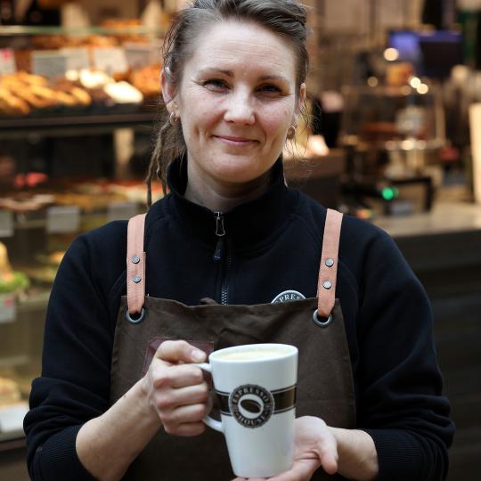 En smilende Espresso House-medarbejder står klar til at servere en lækker kop kaffe. Koppen er fyldt til randen med dampende kærlighed.